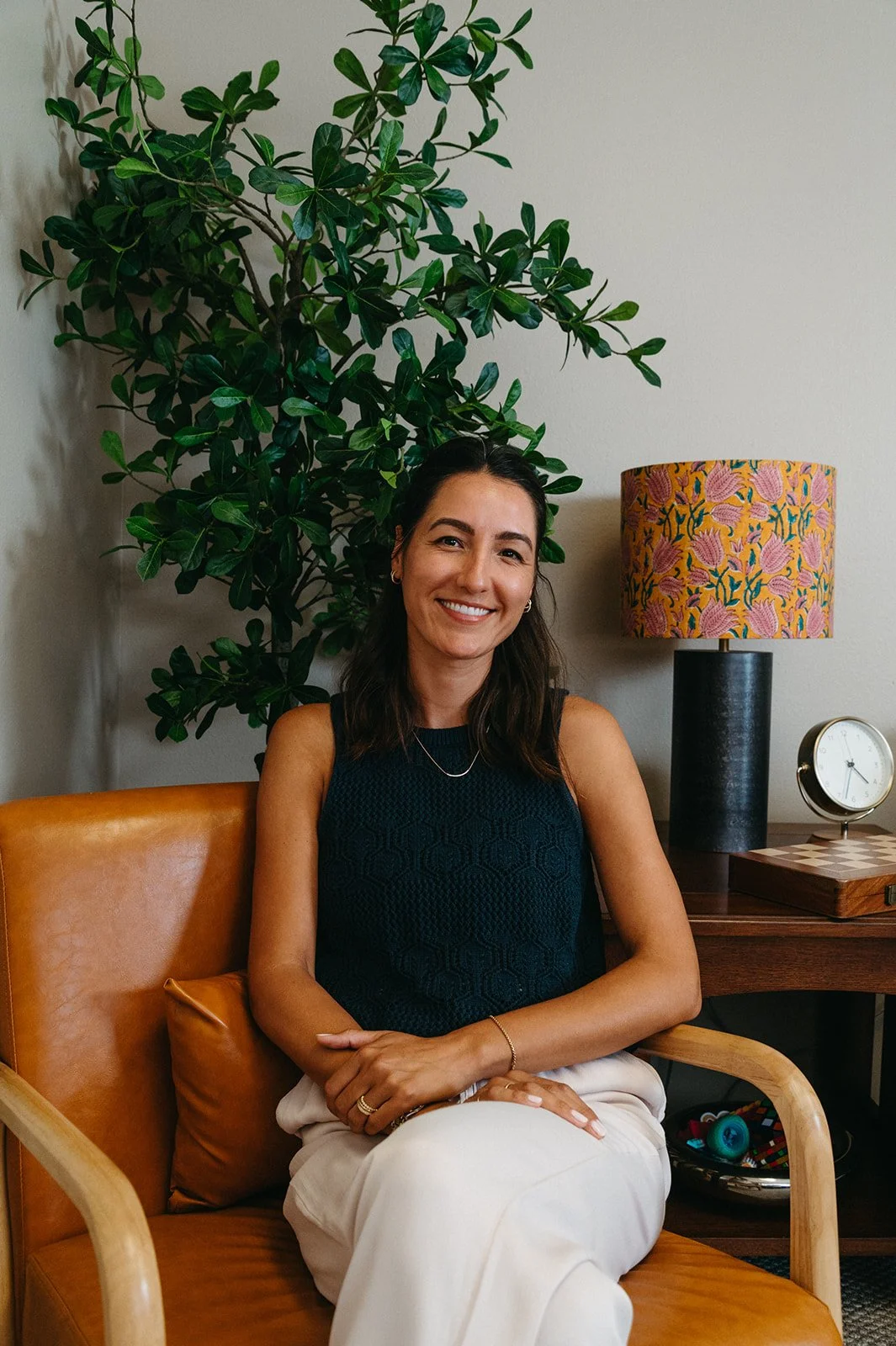 A woman with dark brown hair and a smile, sitting on a tan leather chair with a cushion, in a room with a large green plant behind her, a wooden side table with a colorful lampshade, a clock, and a tray with a small toy.