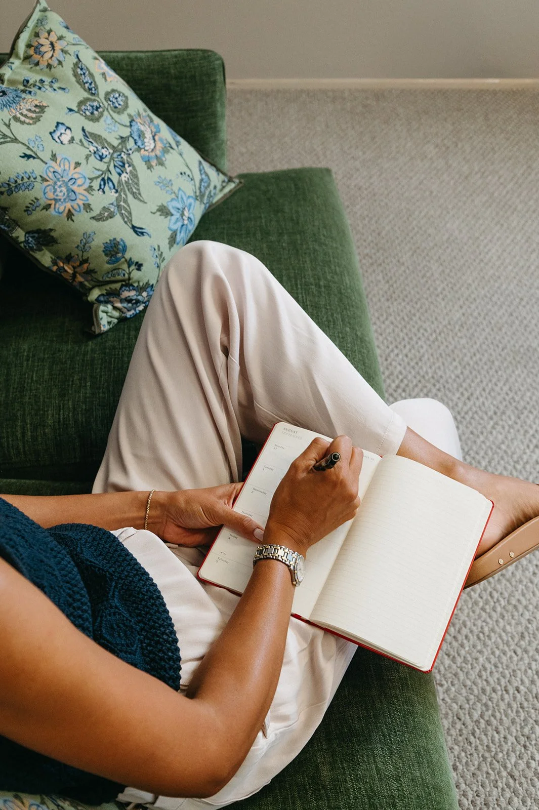 Person sitting on a green sofa, writing in a notebook with a pen. The person is wearing a navy blue top and white pants, with a watch and bracelet on their left wrist. There is a decorative pillow with floral patterns on the sofa. The setting appears to be a cozy indoor space with carpeted flooring.