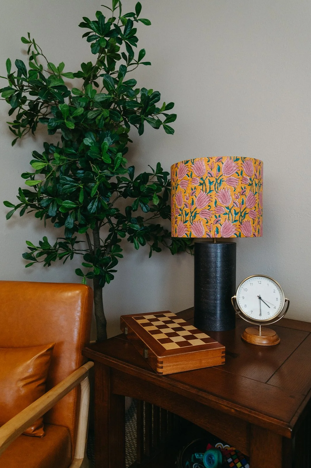 Wooden table with a floral lampshade, a vintage clock, and a chessboard, next to a leather armchair and a large green houseplant against a beige wall.