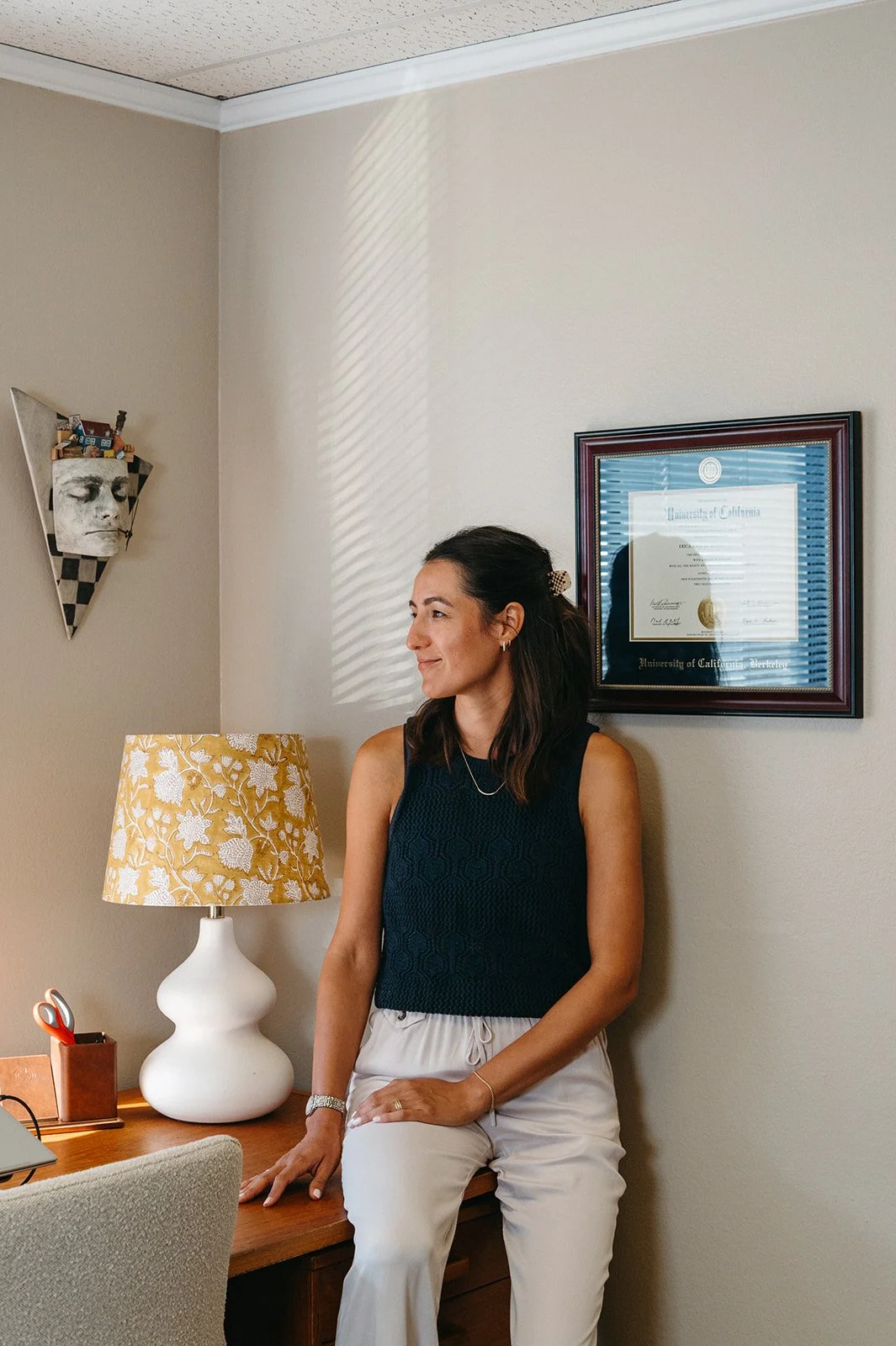 A woman with dark hair wearing a black sleeveless top and white pants, sitting on a wooden desk in a beige-walled room, with a framed diploma from the University of California, Berkeley, a yellow floral lampshade, and a modern wall art piece of a fac
