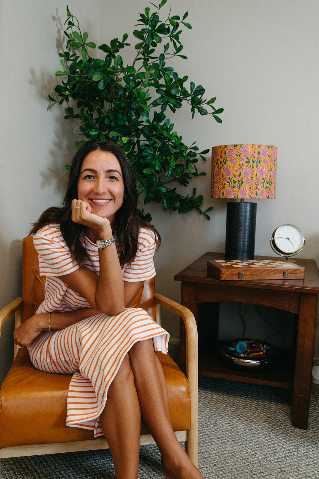 A woman sitting in a wooden chair with a caramel leather seat, smiling, with a houseplant, a lamp with a floral shade, and a clock on a wooden side table behind her.