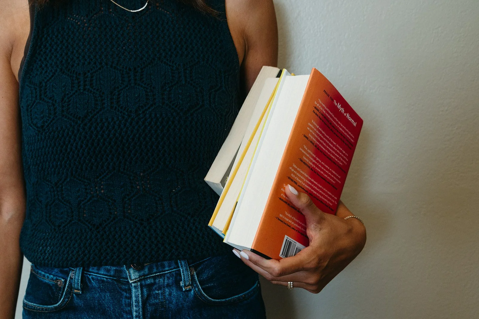 A person wearing a black, sleeveless, woven top and blue jeans, holding a stack of books against a plain wall. The person's left hand is holding the books, which include a red book titled 'The Myth of Normal,' with the cover partially visible.