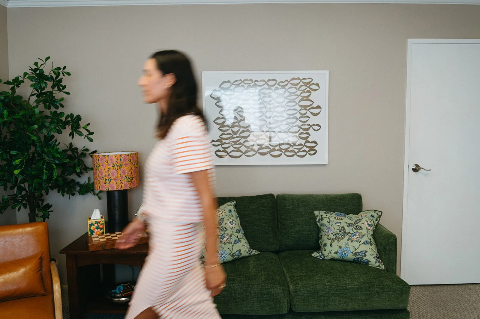A woman in a red and white striped dress walking past a living room with a green couch, decorative pillows, a side table with a tissue box, a colorful lamp, a large leafy plant, and framed wall art.