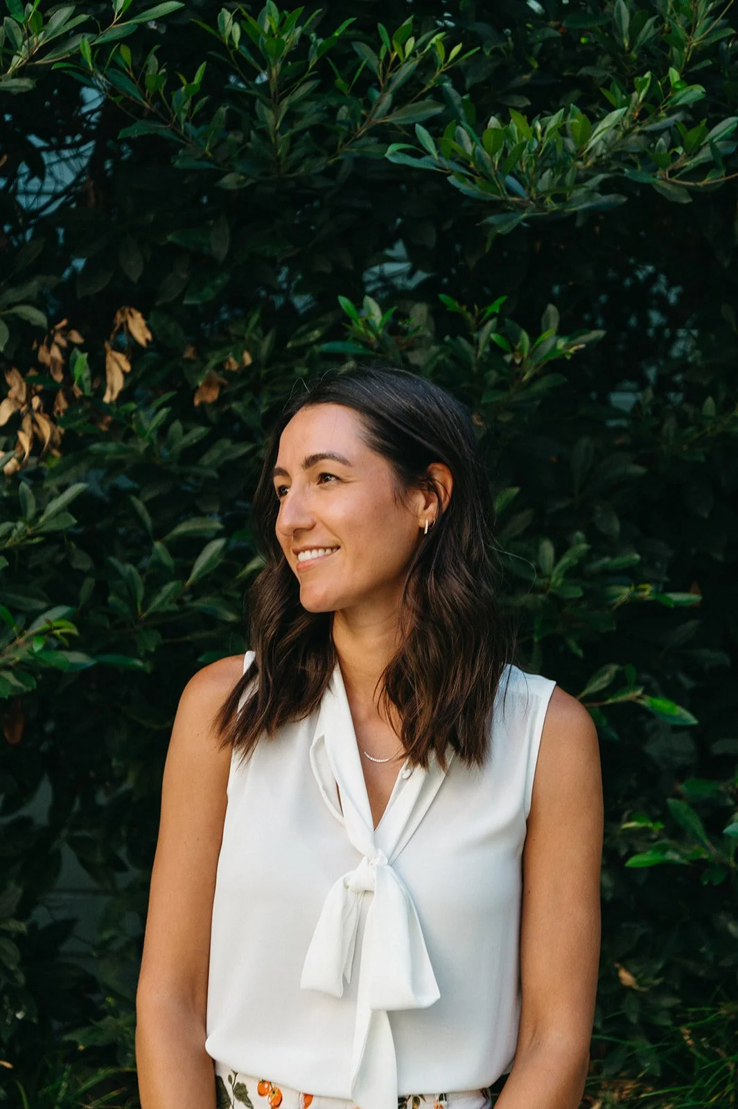 A woman with dark brown wavy hair smiling and looking to the side, standing in front of a leafy green hedge, wearing a white sleeveless top with a bow tie.