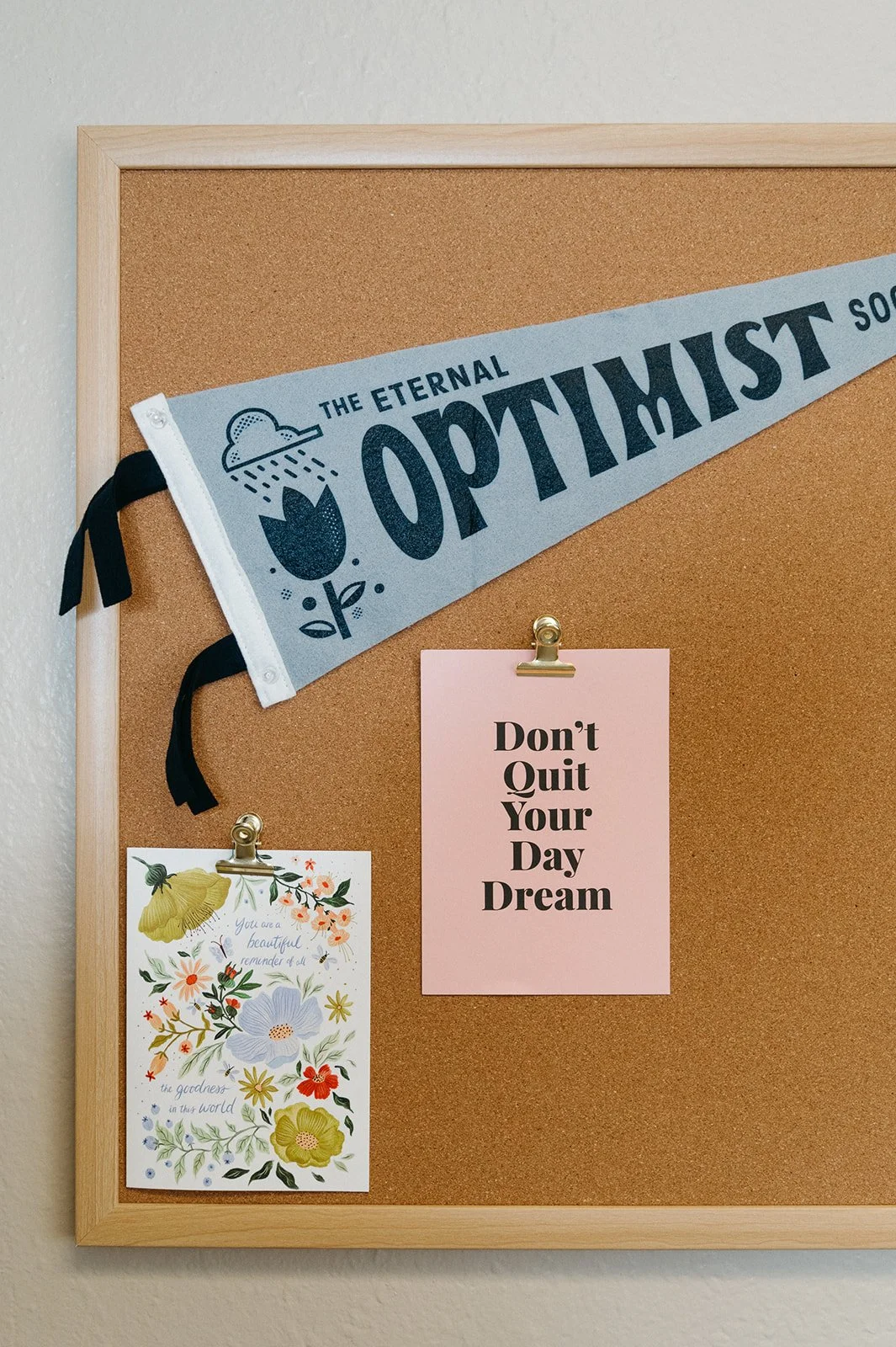 A cork bulletin board with a light wood frame, decorated with motivational and floral notes, and a blue banner with black text reading "The Eternal Optimist".