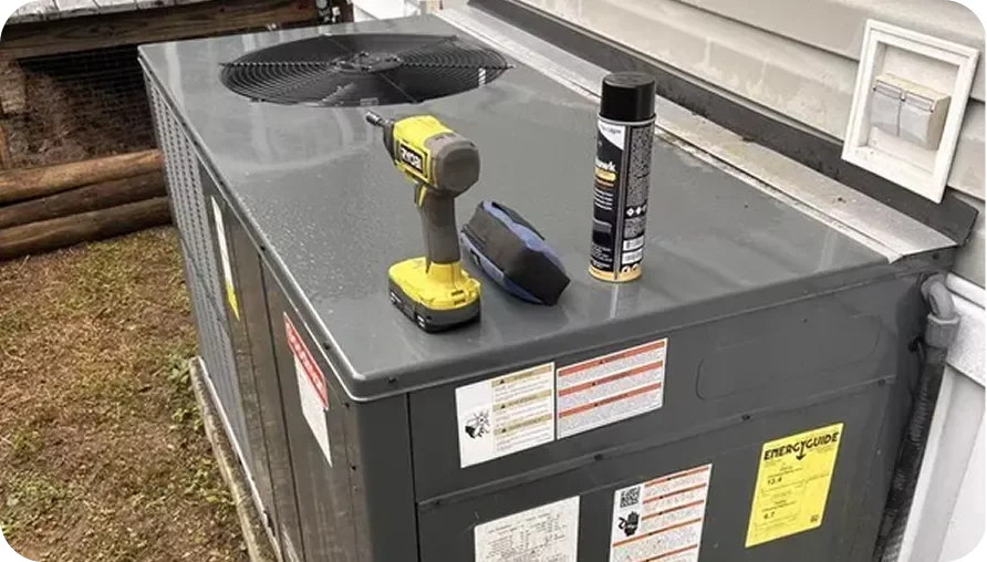 A gray outdoor air conditioning unit with a yellow drill, a spray can, a small bag, and a black object on top.