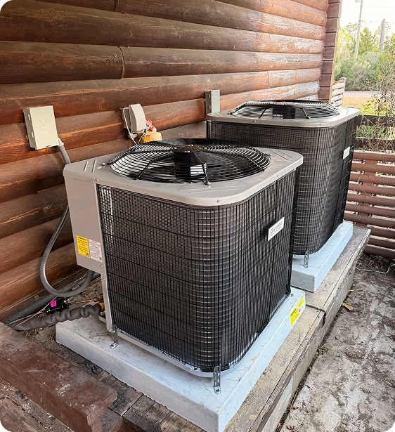 Two outdoor air conditioning units on concrete pads beside a wooden wall, with electrical wiring and cables connected.