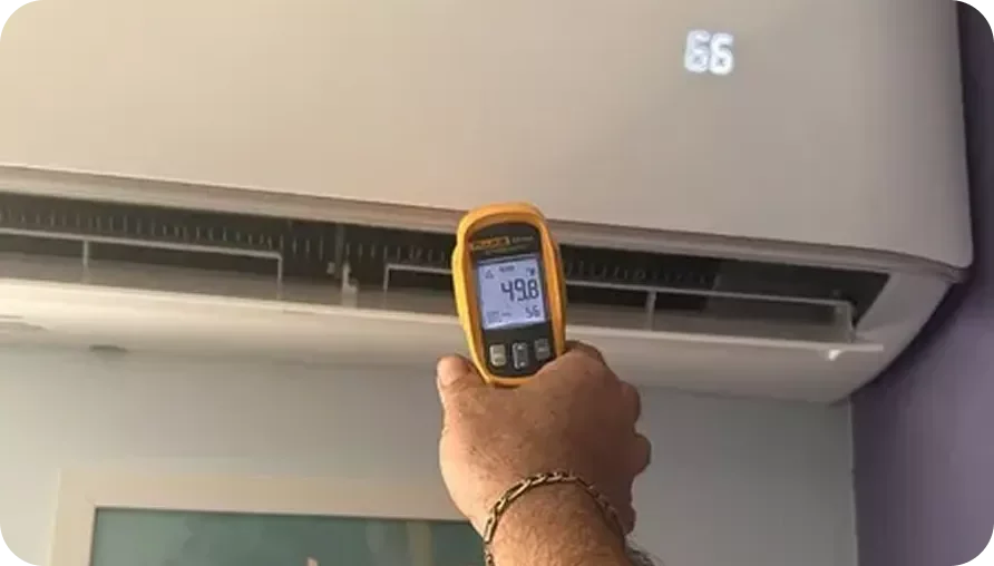 A person holding a digital thermometer measuring the temperature of an air conditioning unit, displaying 49.8°F.