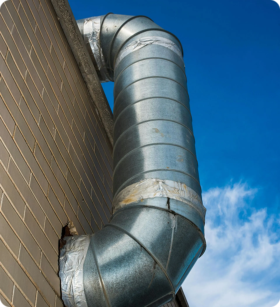 A large industrial metal exhaust vent pipe protruding from a brick building against a blue sky with some clouds.