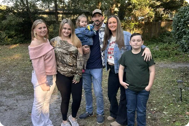 Group of six people outdoors, including two young girls, one toddler, one teenage girl, one teenage boy, and an adult man, standing on a dirt path with trees and a wooden fence in the background.