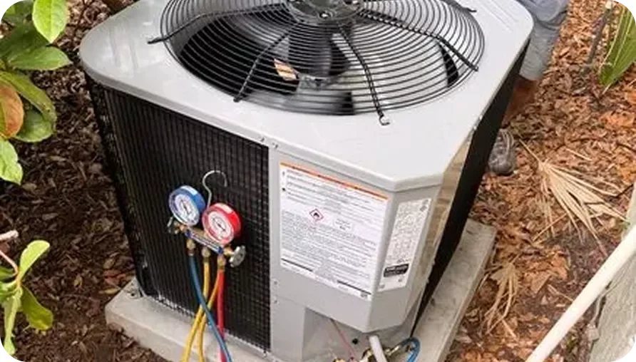 An outdoor air conditioning unit with refrigerant gauges attached, placed on a concrete pad surrounded by plants and mulch.
