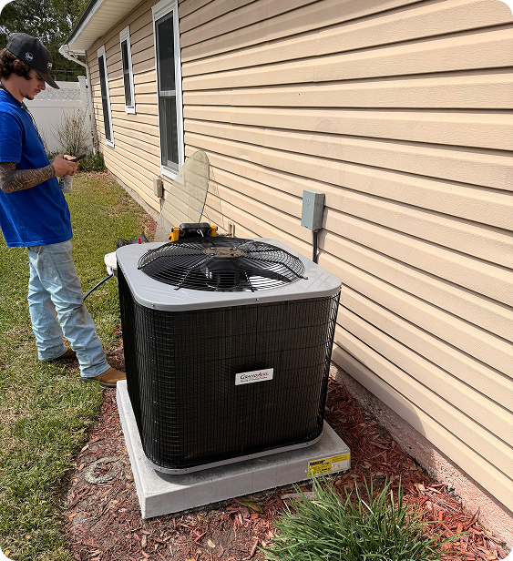A man in a blue shirt and jeans standing outside next to an air conditioning unit installed on a concrete pad beside a beige house with siding. The man appears to be working or inspecting the unit, which has a black grill on top and a fan. The backyard has grass and mulch with plants near the house.