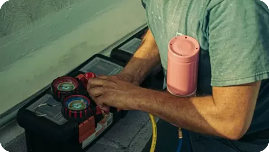 Person with a pink portable speaker, wearing a green t-shirt, working with tools and a yellow hose on a dark surface.