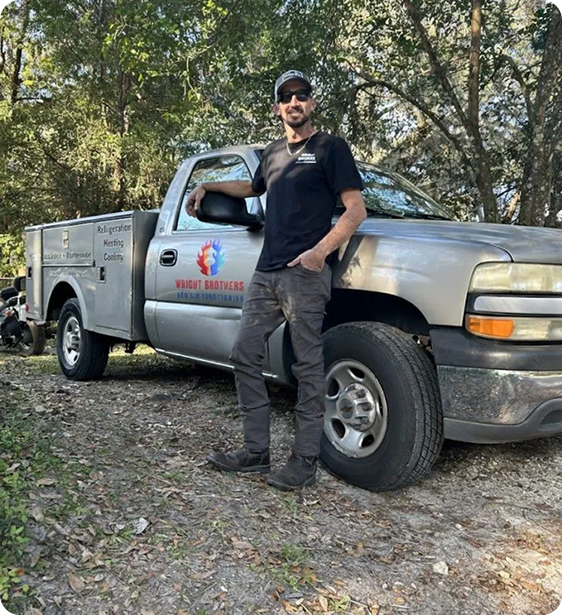 A man with a beard and sunglasses standing next to a gray work truck with a logo that reads 'Weight Brothers' and 'Refrigeration, Heating & Cooling' parked outdoors among trees.