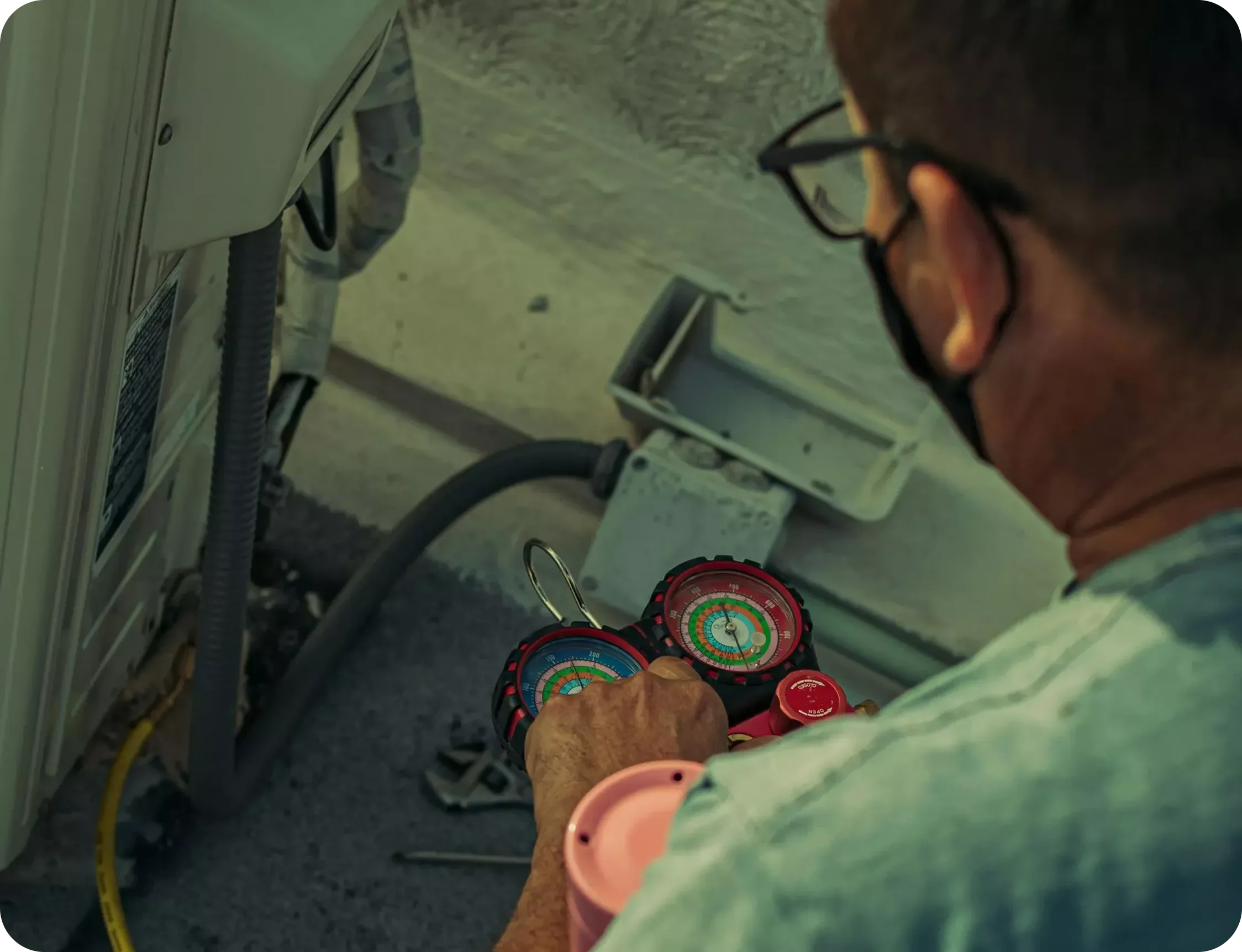 A person with glasses and a mask using hand tools to work on an appliance or machine, with gauges and hoses nearby.