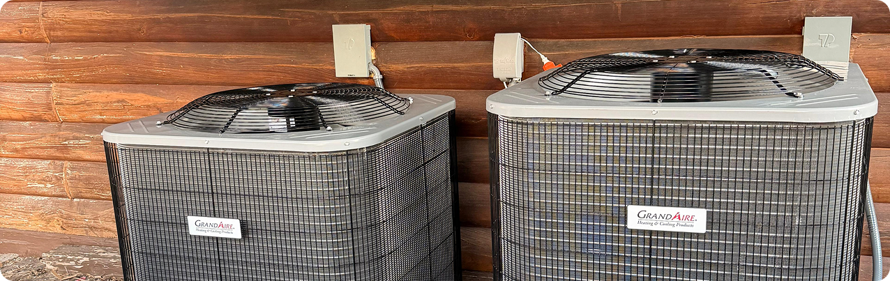 Two air conditioning units outside against a wooden wall with electrical cords plugged into outlets.