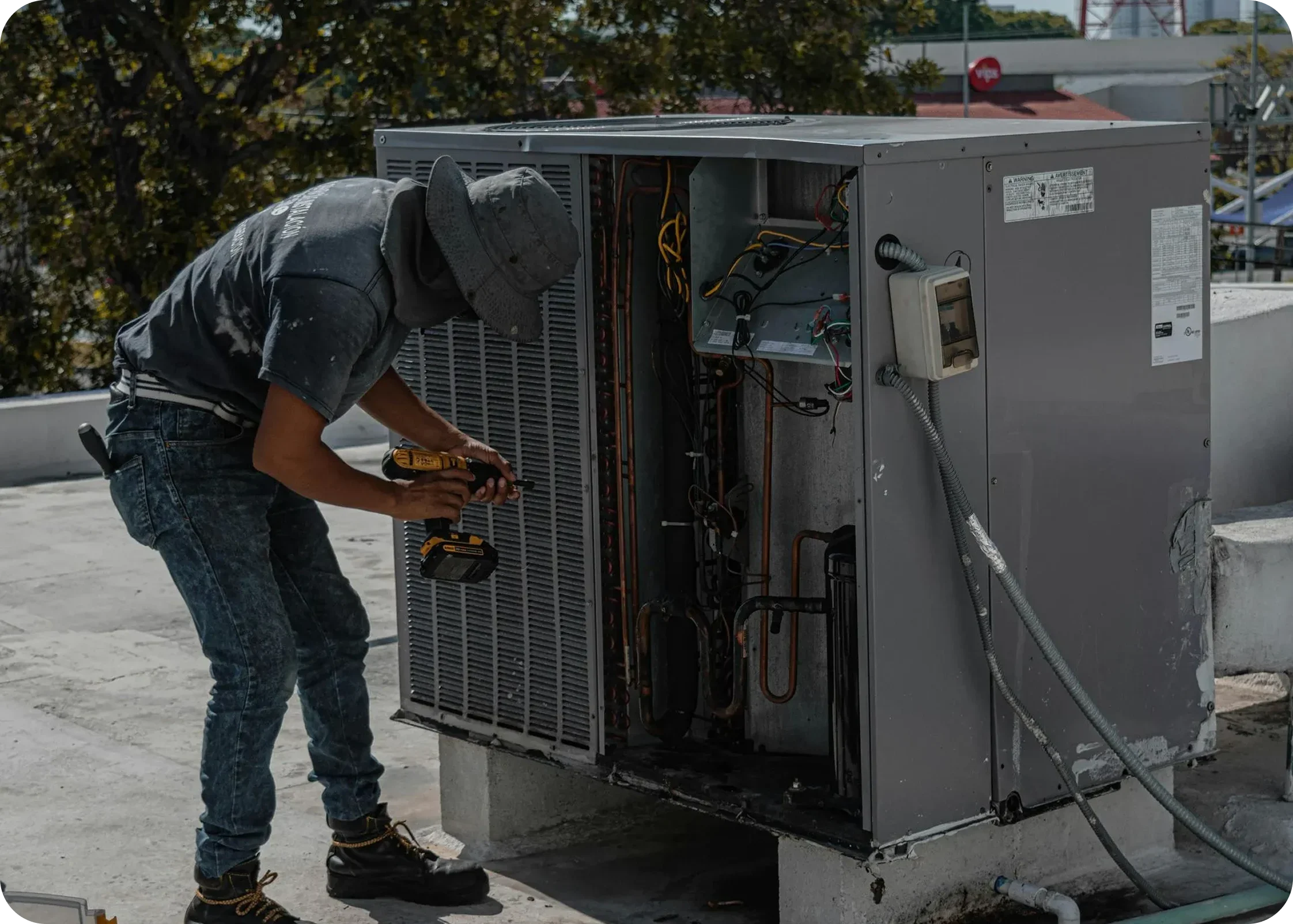 A person working on an outdoor air conditioning unit with tools, wearing a hat and gloves, with trees and buildings in the background.