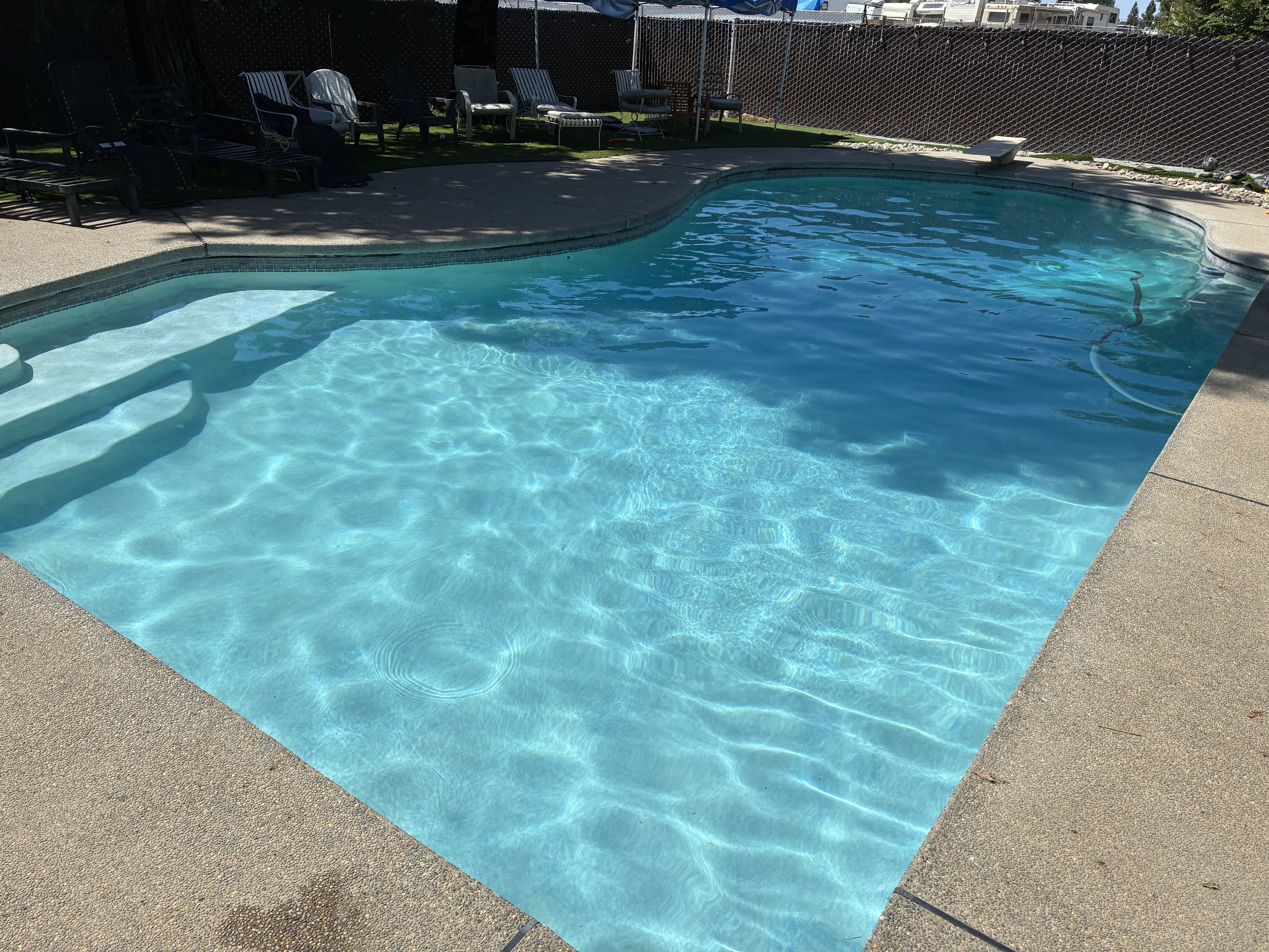 A backyard swimming pool with light blue water, surrounded by a tan concrete pool deck. Several black and white lounge chairs and patio furniture are placed along the back side of the pool, with a tall wooden fence enclosing the area. Shadows from nearby trees are cast on the deck and the water surface.