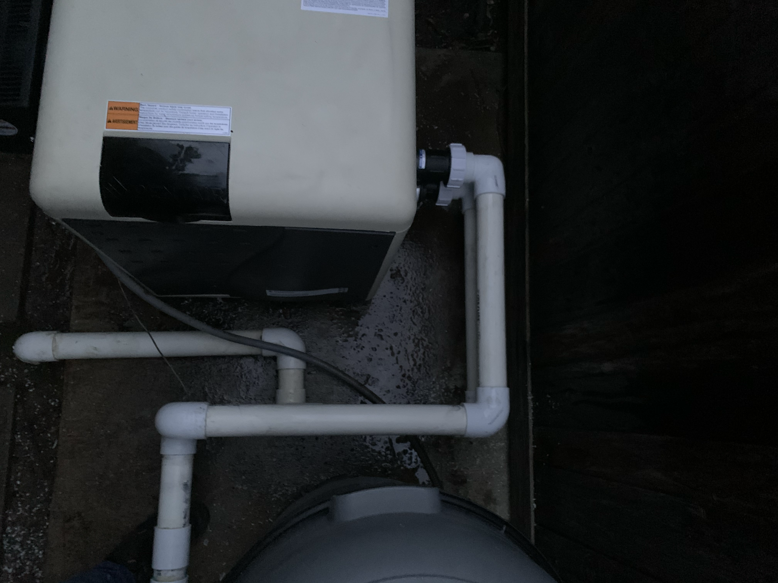 Image of an outdoor water pump or filtration system, surrounded by white PVC pipes and fittings, installed on a concrete or soil surface.