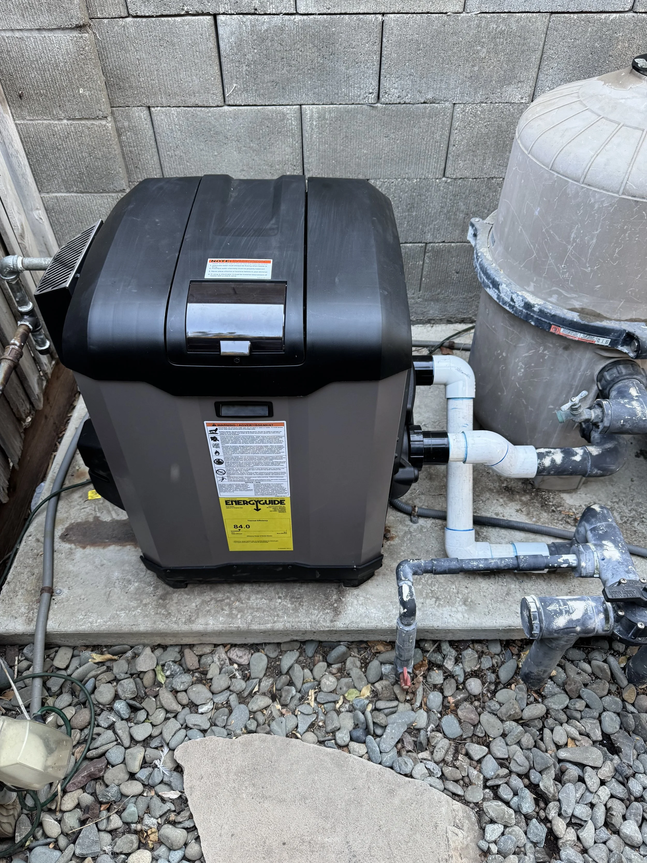 An outdoor scene showing a black pool filter pump on a concrete slab, with attached white PVC pipes, a large beige sand filter, and a bed of small gray rocks surrounding the setup. A gray cinder block wall is in the background.