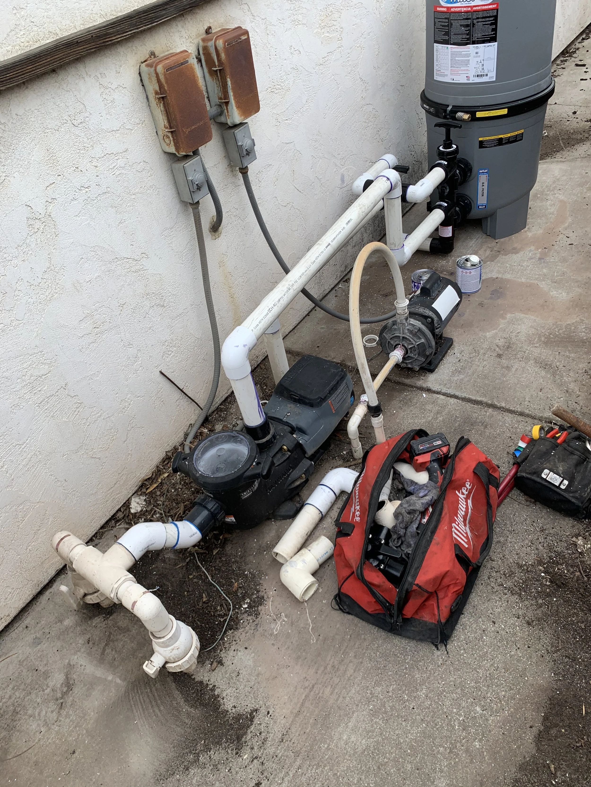 A plumbing system installation on an exterior wall, including PVC pipes, a water filter, an electric pump, and a red Milwaukee tool bag containing plumbing tools.