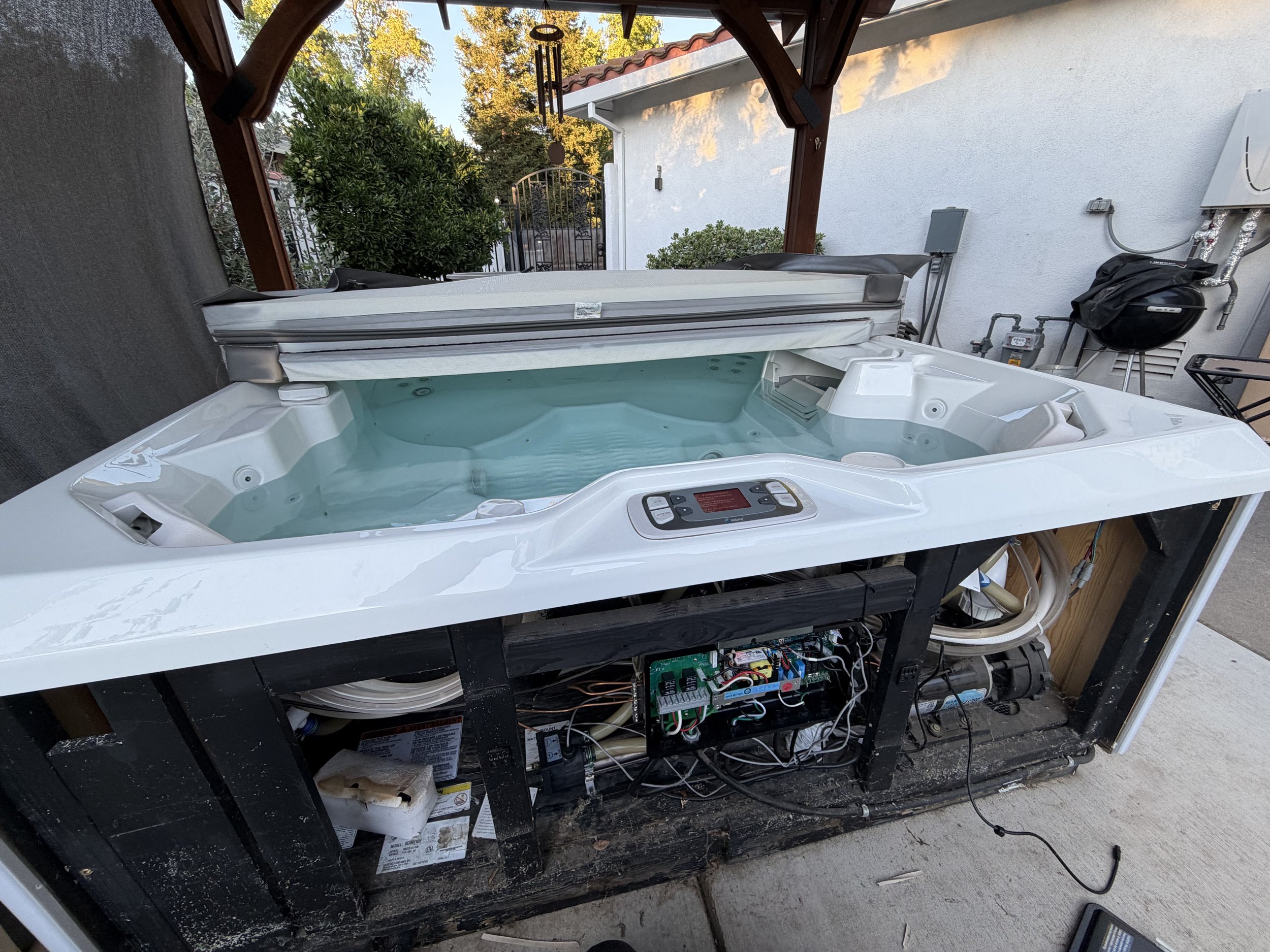 An outdoor hot tub or Jacuzzi with its electronic control panel visible, under a wooden pergola with a wind chime hanging from it. The background shows a white wall with pipes and electrical connections, and a lush green shrub.