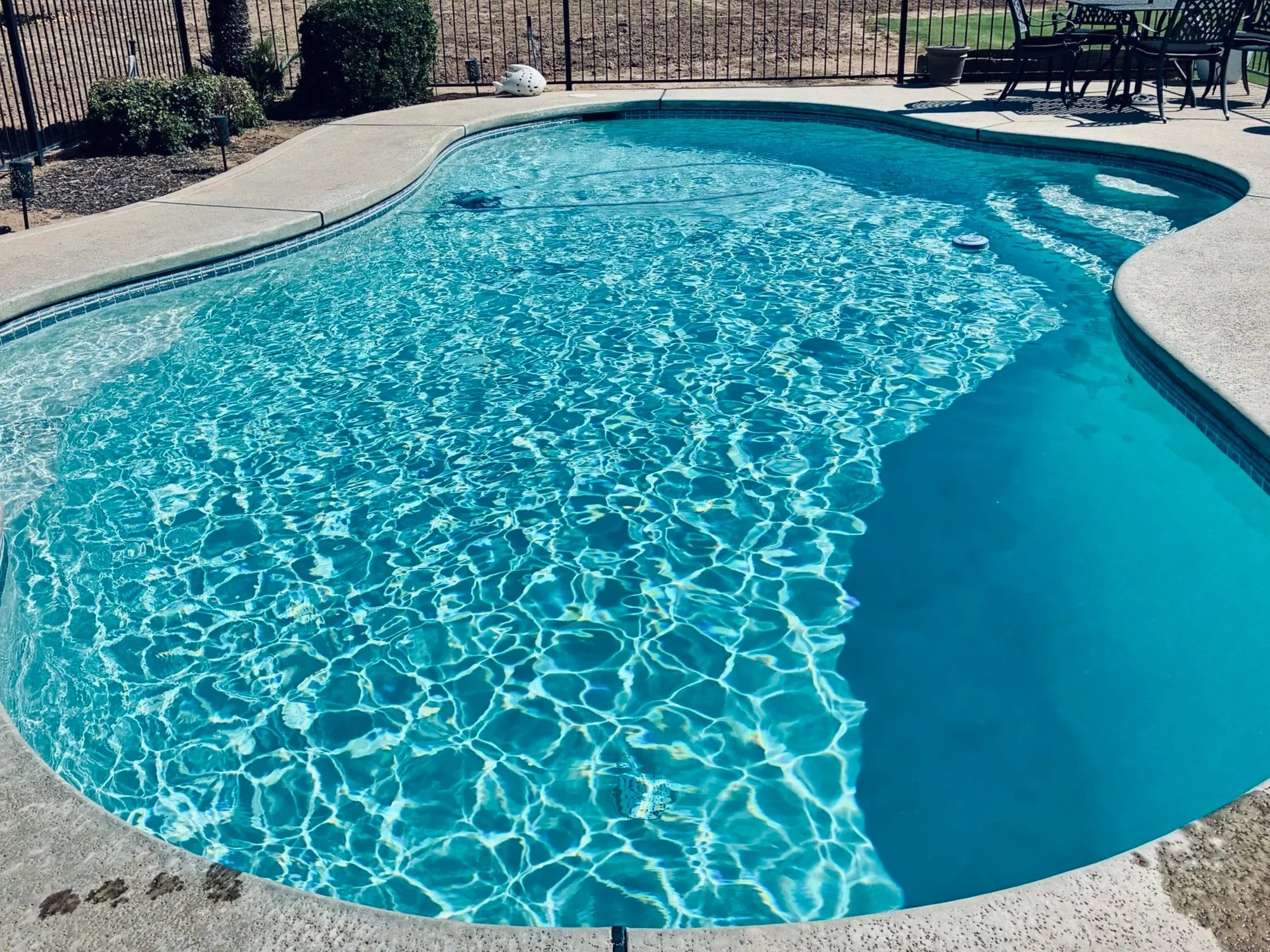 Residential backyard with a blue in-ground swimming pool, patio furniture in the background, and a fenced yard.