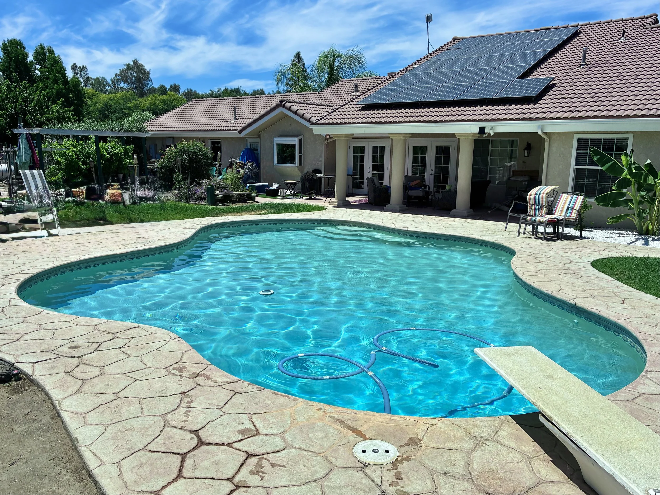 A backyard with a swimming pool, patio chairs, and a house with a tiled roof and solar panels.