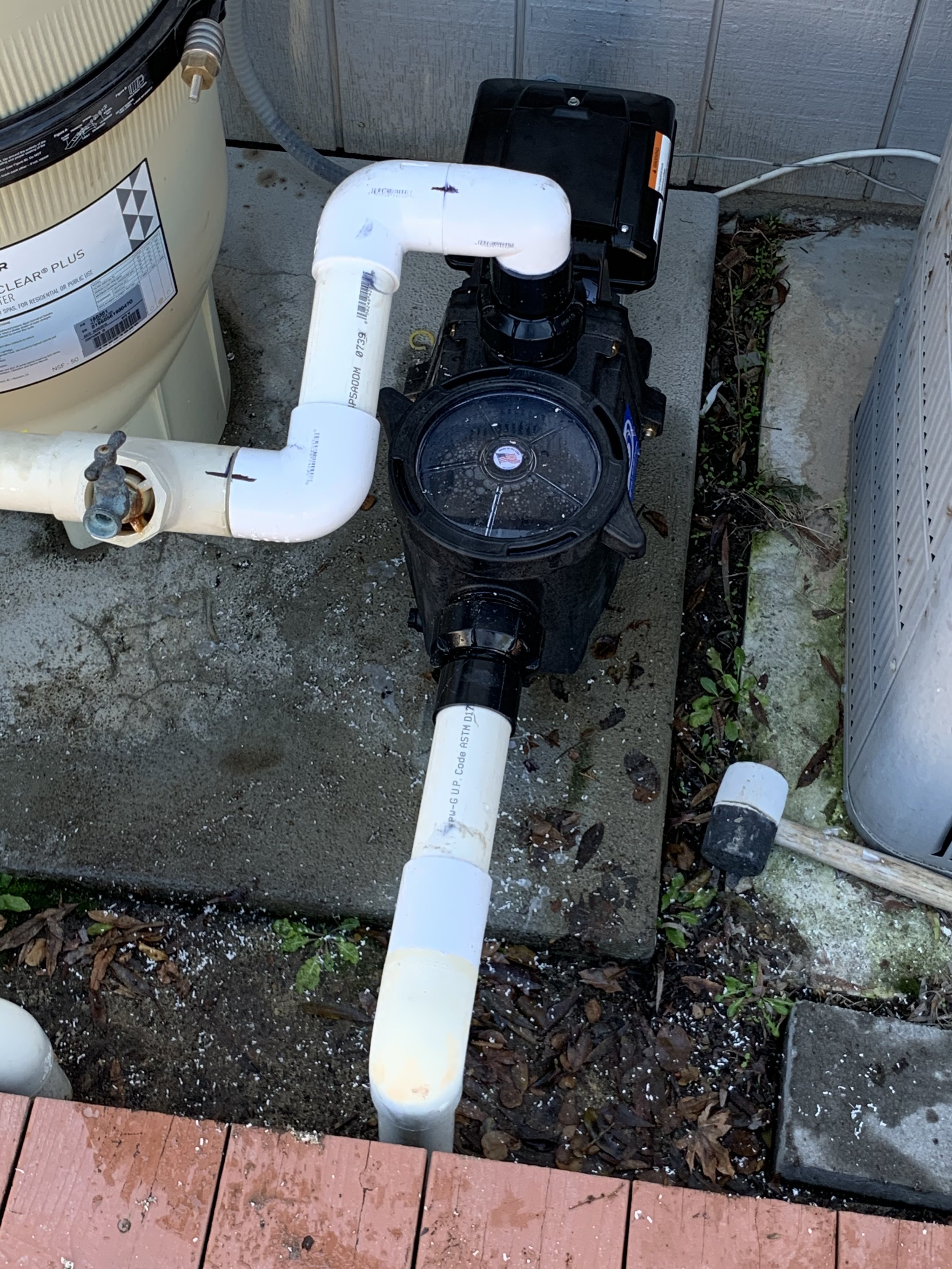 The image shows an outdoor area with a pool pump and filter system connected to PVC pipes. There is some dirt, leaves, and foliage on the ground, along with a section of a red wooden deck at the bottom.