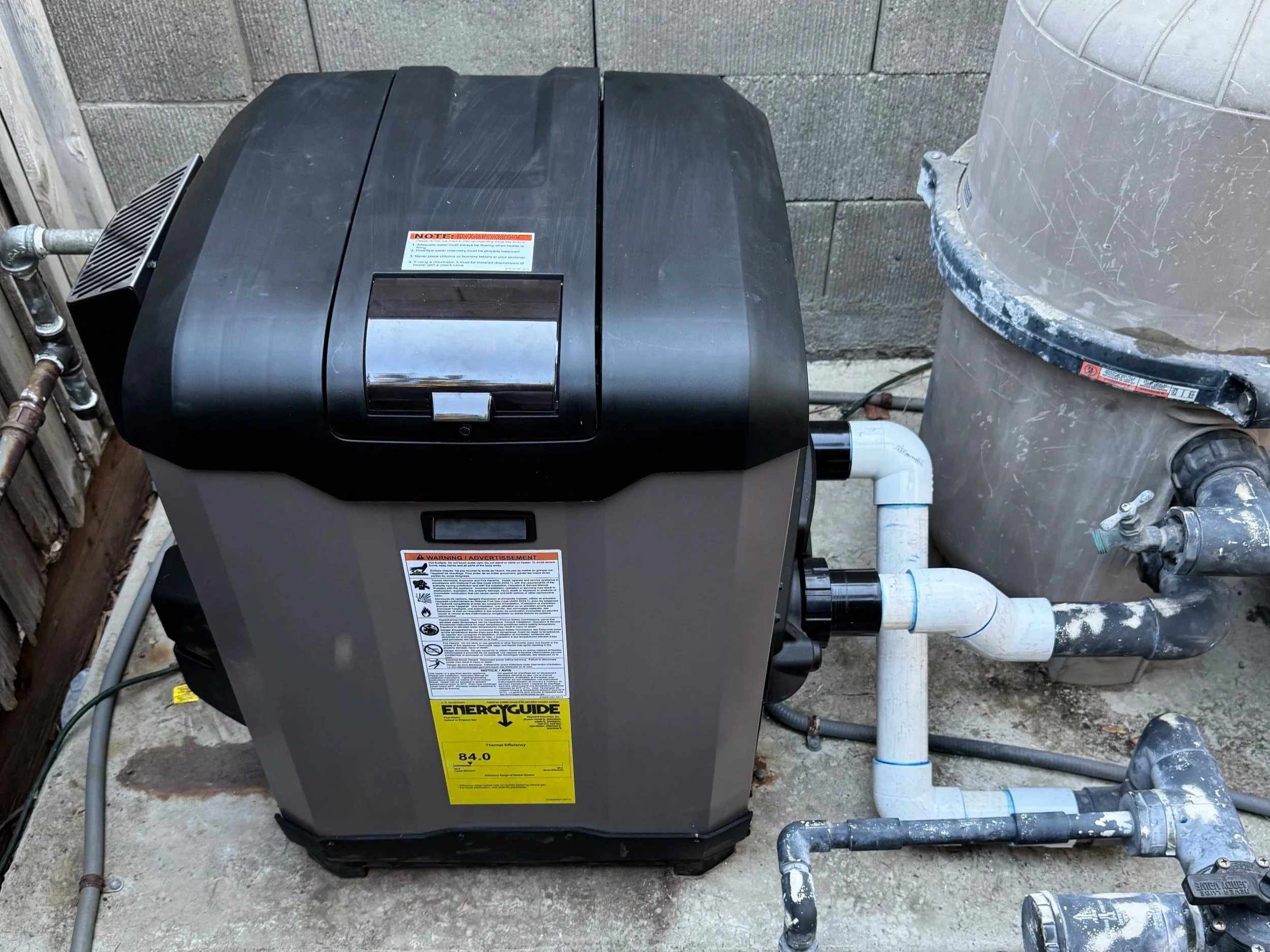 Hayward pool heater installed next to a cinder block wall, with PVC pipes and metal fittings connected to it.