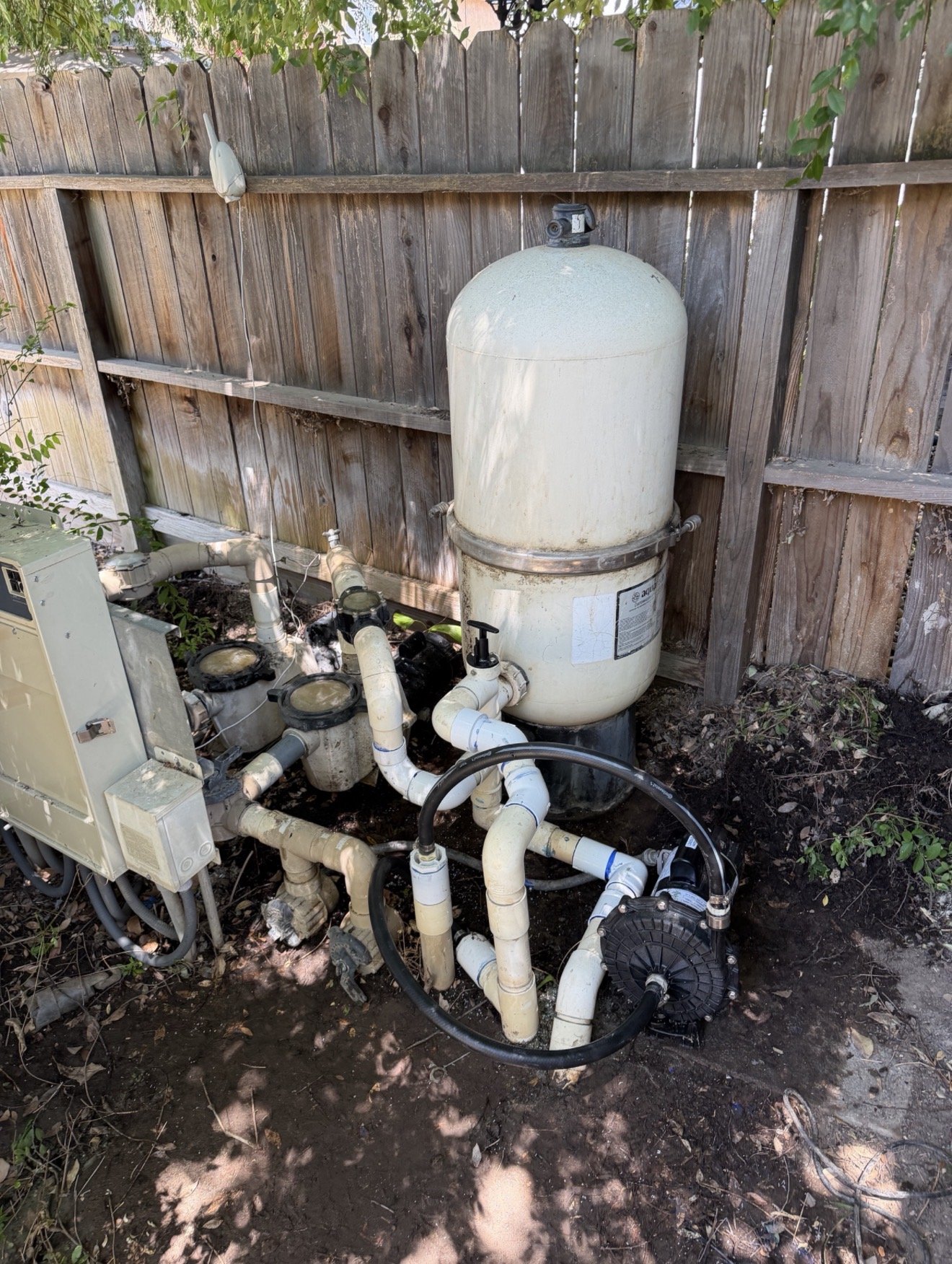 An above-ground swimming pool pump and filtration system with white pipes connected, set against a wooden fence, on a soil ground with scattered leaves and greenery around.