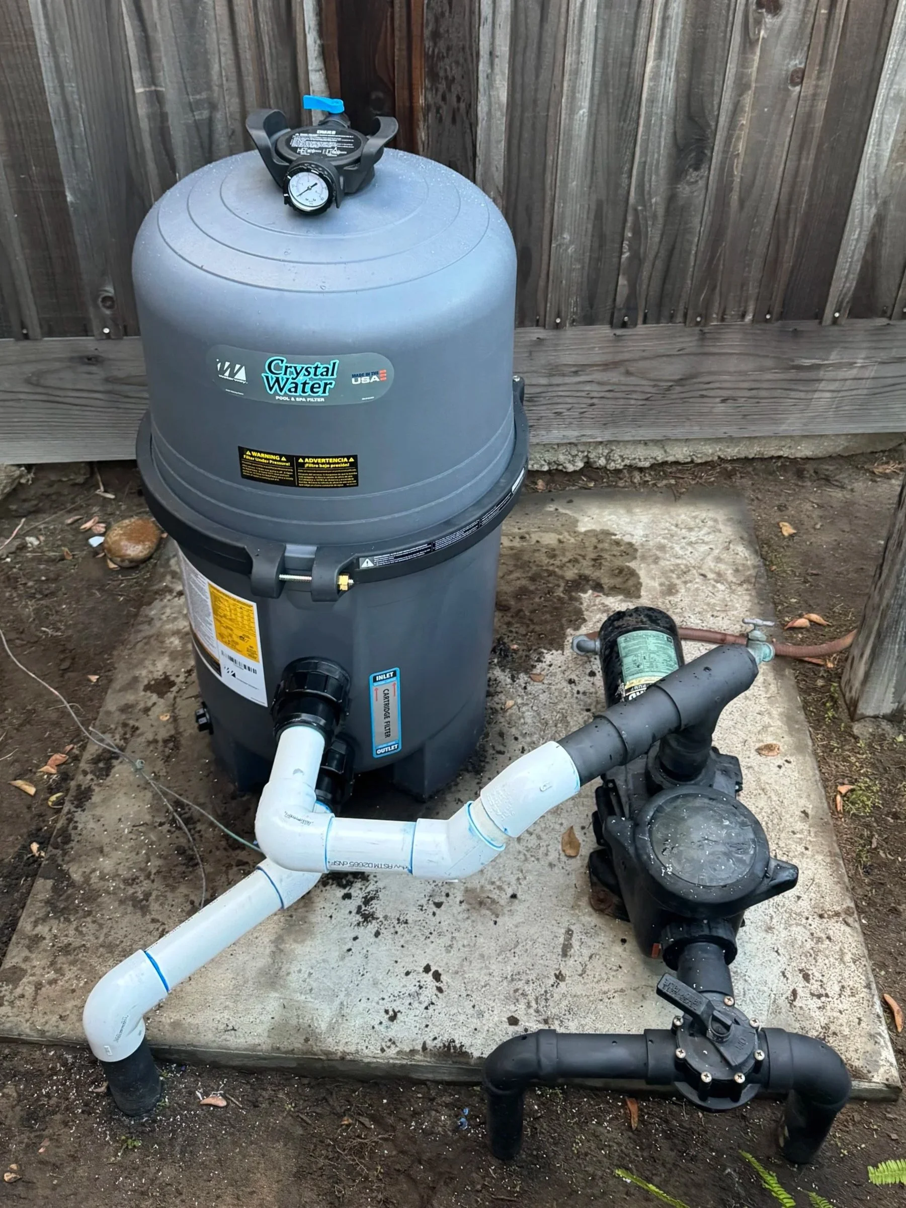 An outdoor pool pump system with pipes, a control valve, and gauges next to a wooden fence.