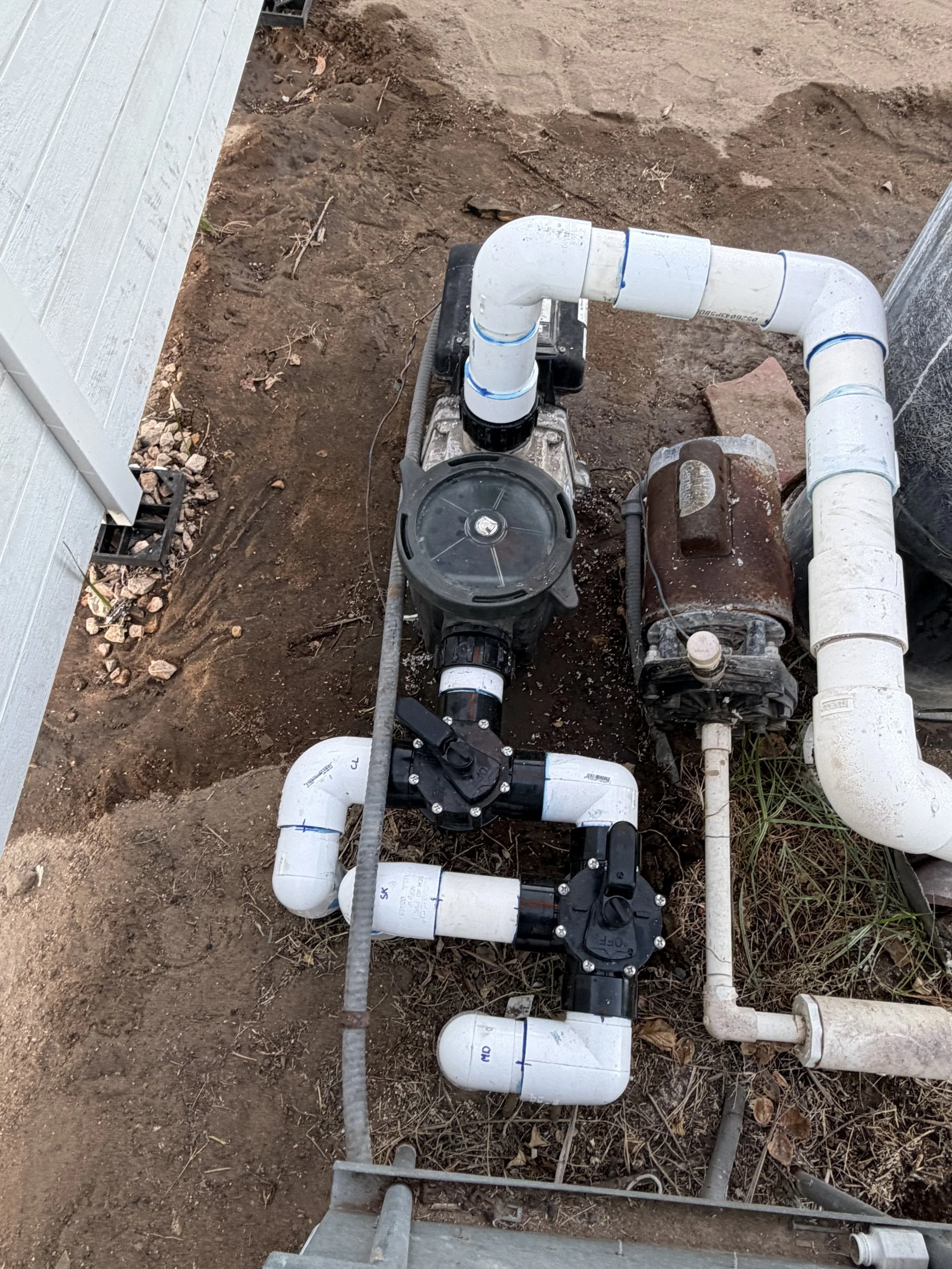 Pipeline system with white pipes, valves, and a pump on dirt ground outside.