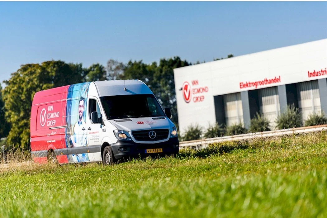 A white delivery van with branding for Van Eijkmond Groep, parked on grass near a building with similar branding, surrounded by green trees.