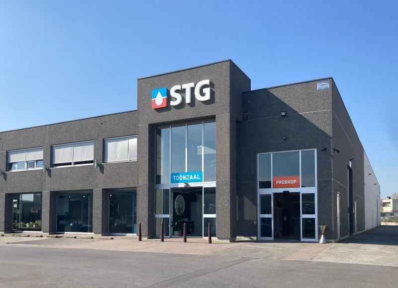 A gray commercial building with the logo 'STG' and a drop symbol, featuring large windows and two entrances labeled 'TOON2AAL' and 'PROSHOP,' against a clear blue sky.