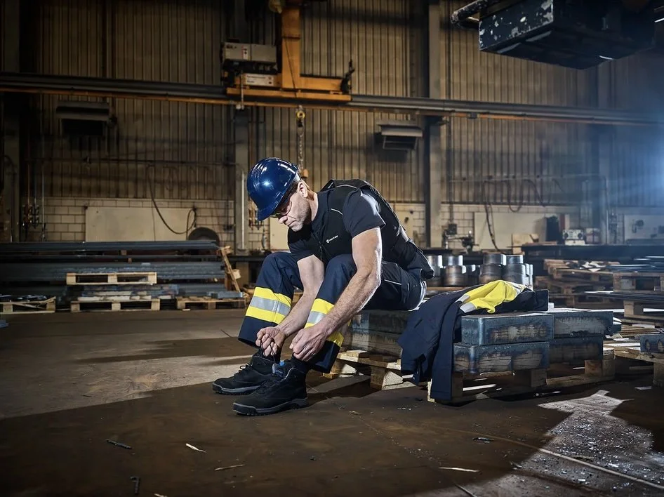 A worker in safety gear, including a blue helmet, is sitting on a wooden pallet in an industrial warehouse, tying his shoelaces with other equipment and pallets visible in the background.