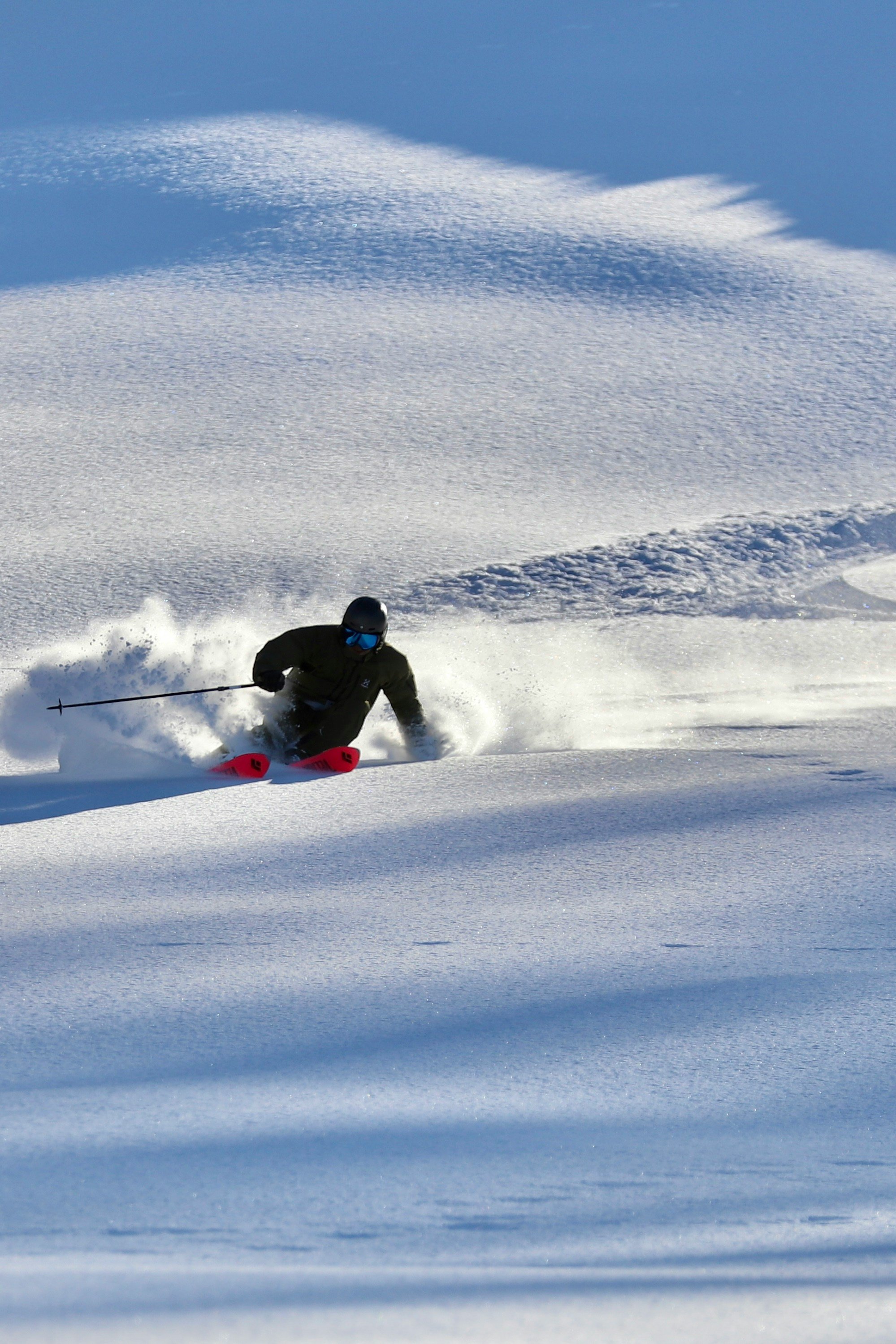 A skier in black gear with blue goggles skiing down a snowy slope, kicking up snow in a powder trail with snow-covered terrain and a clear blue sky in the background.