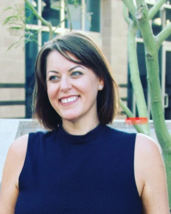 A woman with shoulder-length brown hair, wearing a sleeveless navy blue top, smiling outdoors near a tree.