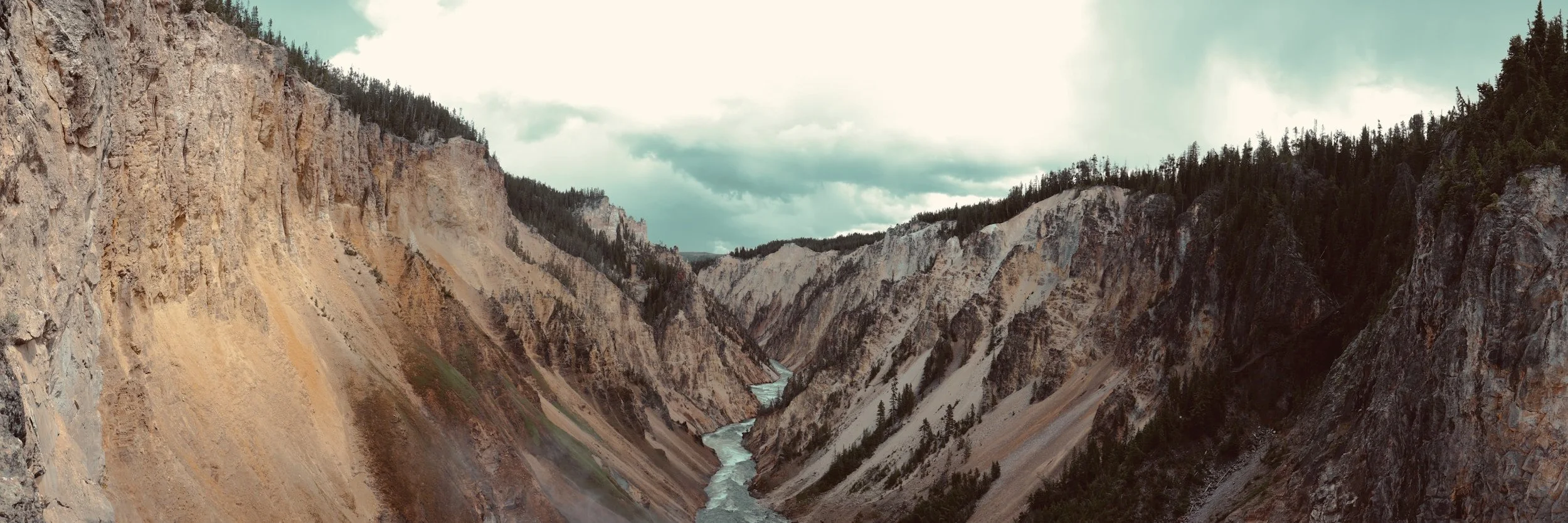 A panoramic view of a deep canyon with steep, rocky walls and a river running through the bottom, surrounded by a forested area under a cloudy sky.