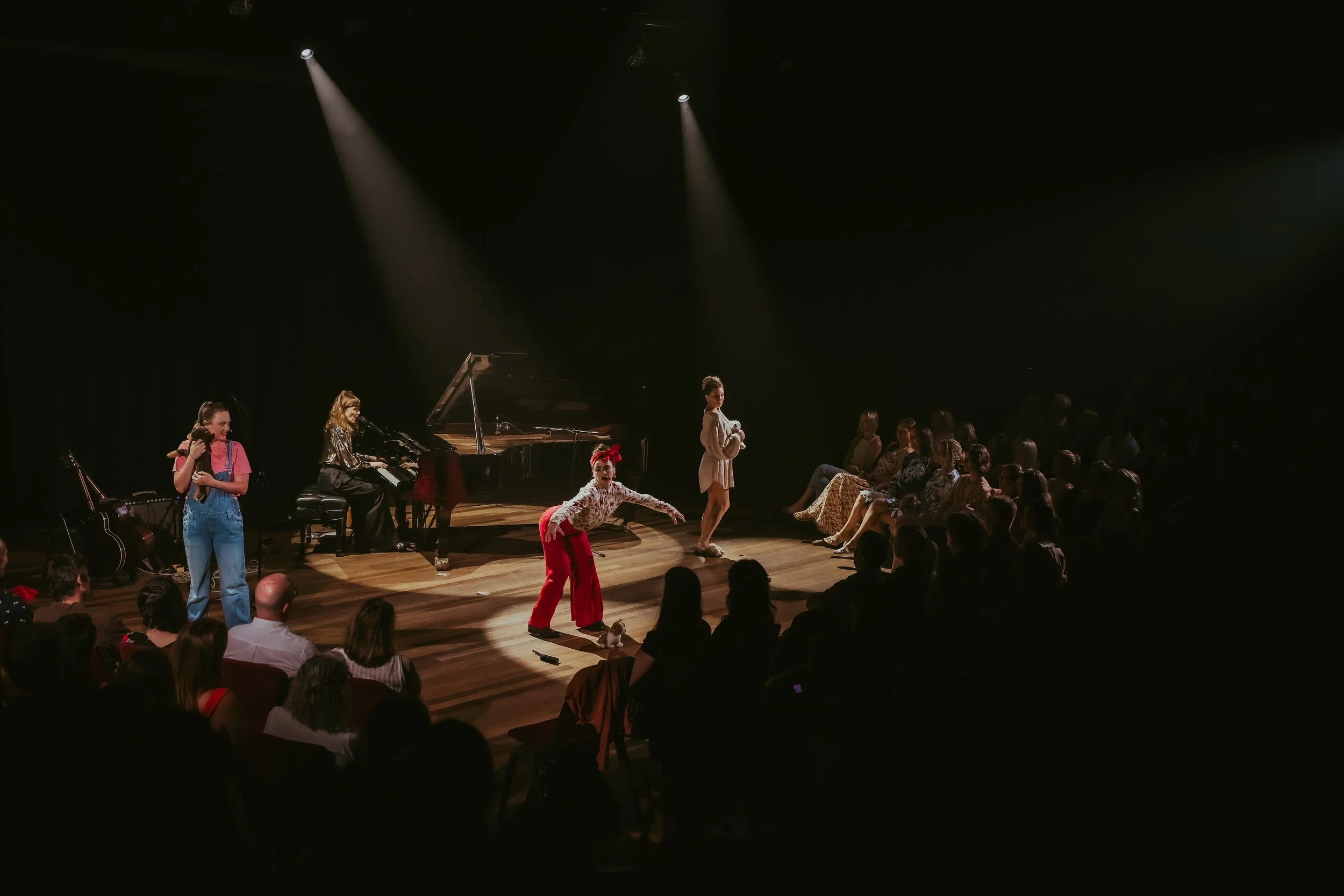 Performers on stage at a concert, including a woman in red pants and a white patterned shirt crouching, another woman in a beige coat standing, a woman playing the piano, and a girl with pigtails listening, with an audience seated in front.