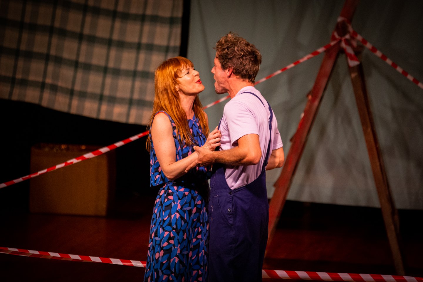 A man and a woman are dancing closely on a stage with a circus tent background and red and white striped tape.