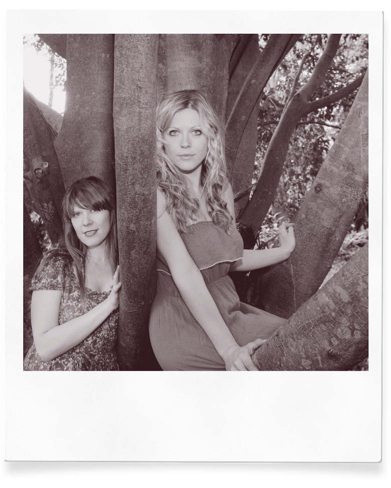 Two young women sitting among tree trunks outdoors, one with long curly hair in a strapless dress, the other with straight hair in a patterned dress, both looking at the camera.