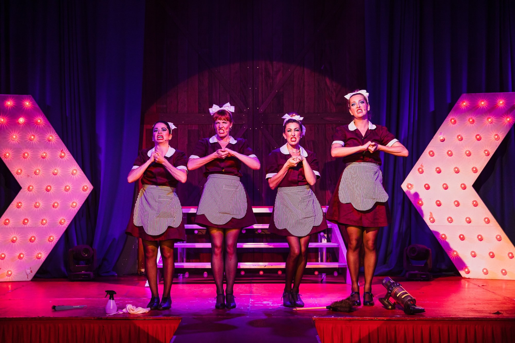 Five women dressed as waitresses stand on a stage with a dark background and large lit-up arrow signs. They are making angry or intense facial expressions, with hands clasped over chests. The stage has props including cleaning supplies and a mop.