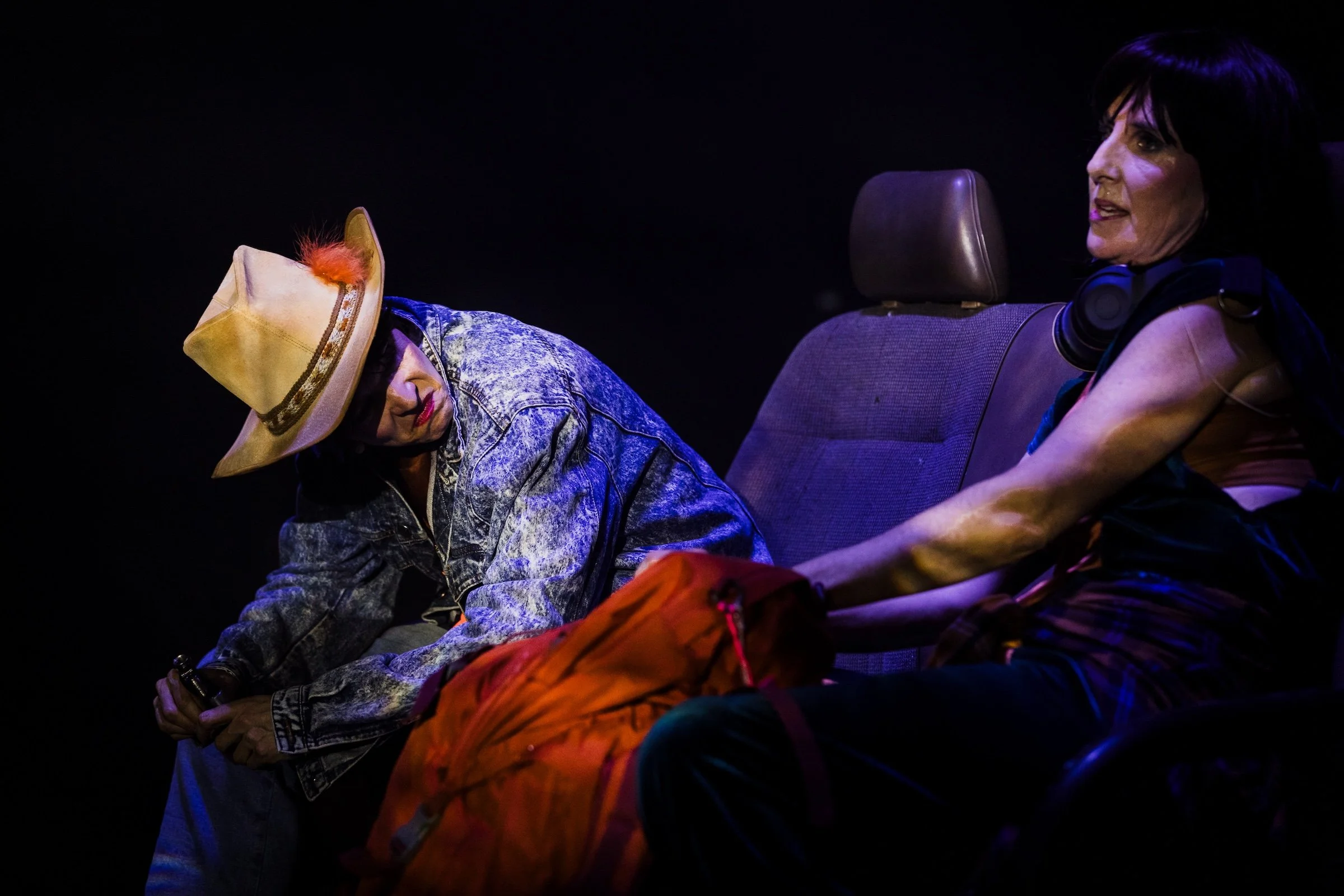 Two women sitting in a dark room; one wearing a cowboy hat with orange feather and denim jacket, the other with short black hair and headphones around her neck.