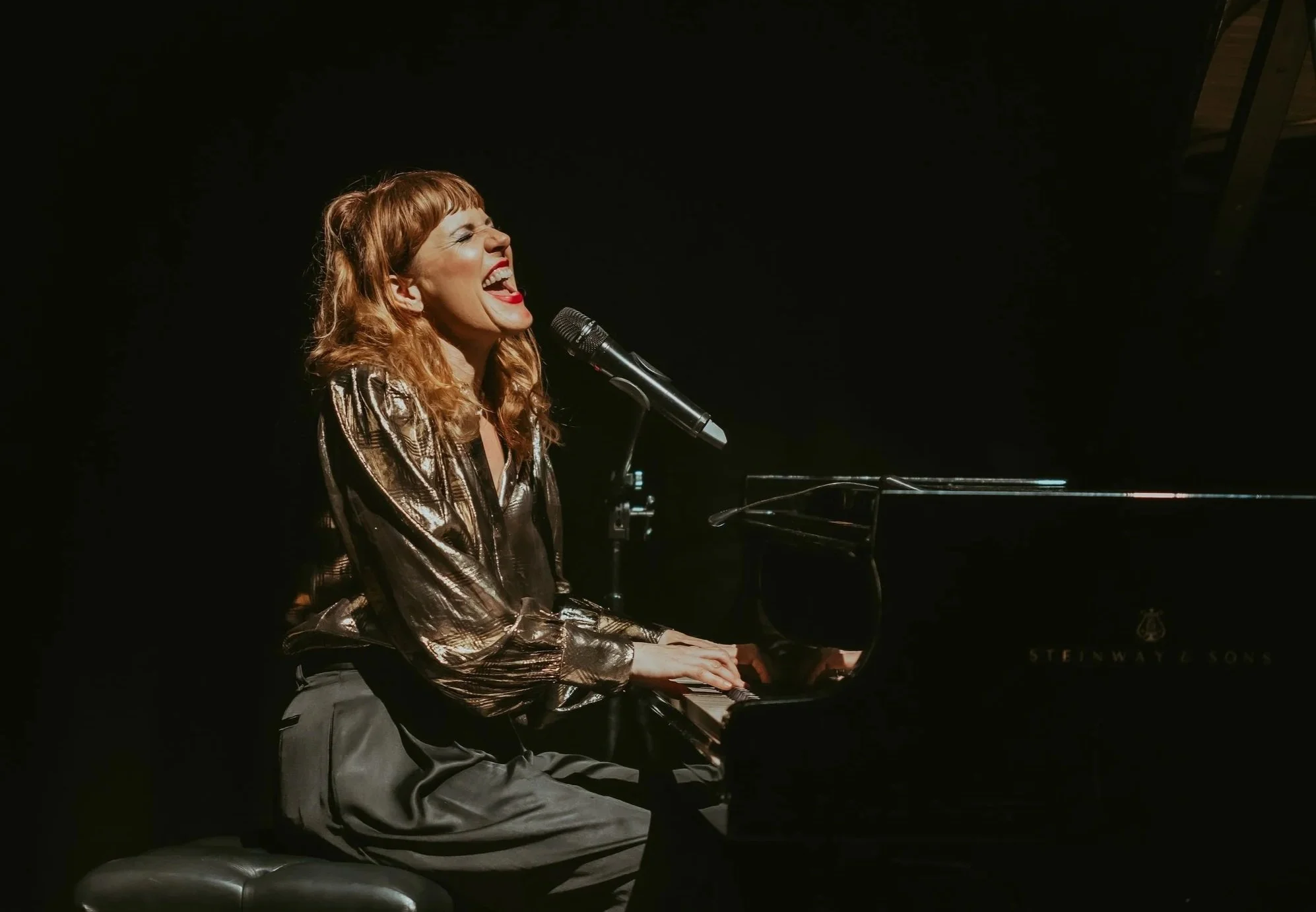 A woman singing passionately while playing a grand piano on stage with a microphone in front of her.