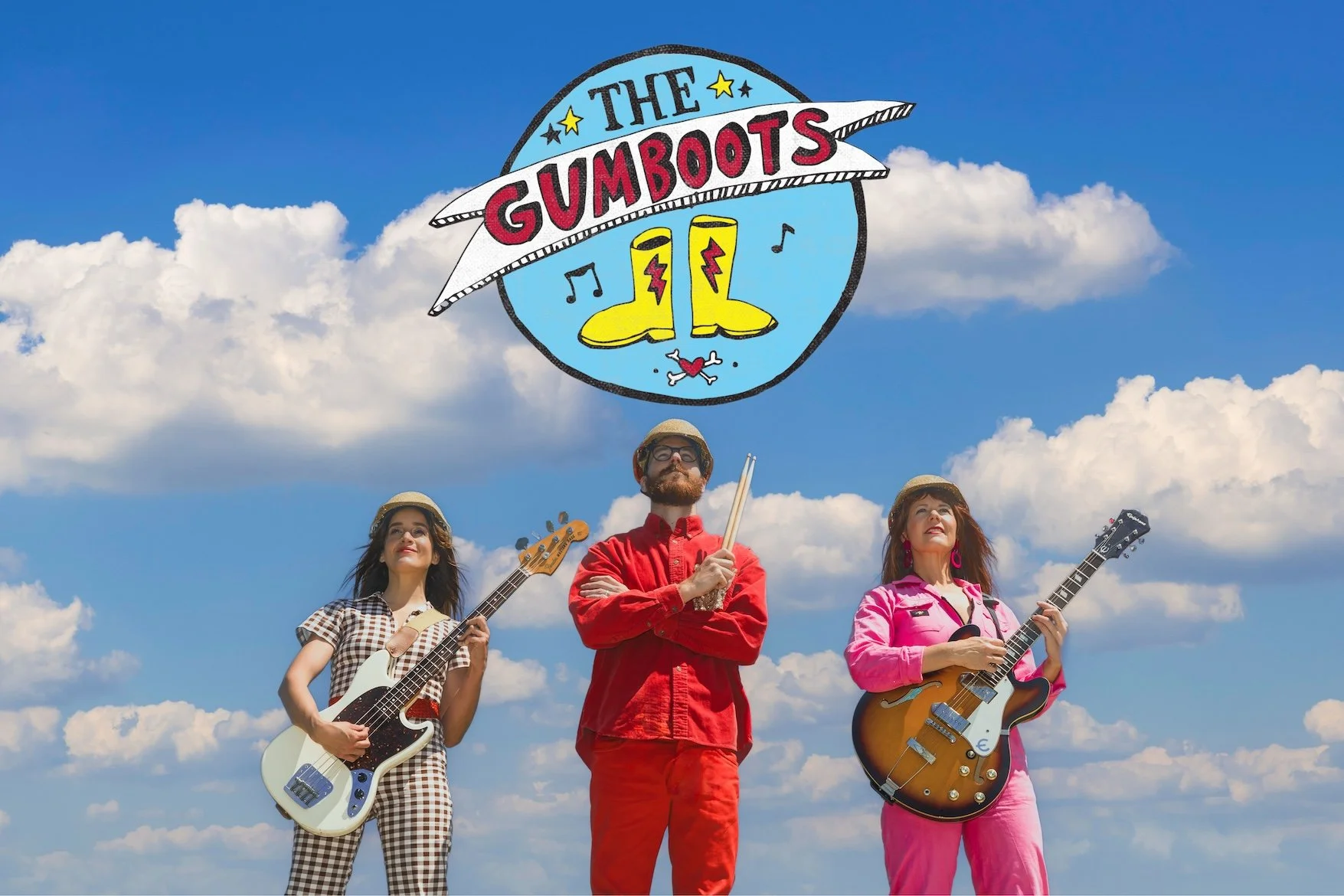 Three members of The Gumboots band outdoors under a blue sky with clouds, holding musical instruments and with a colorful logo above them featuring yellow boots, musical notes, and the band's name.