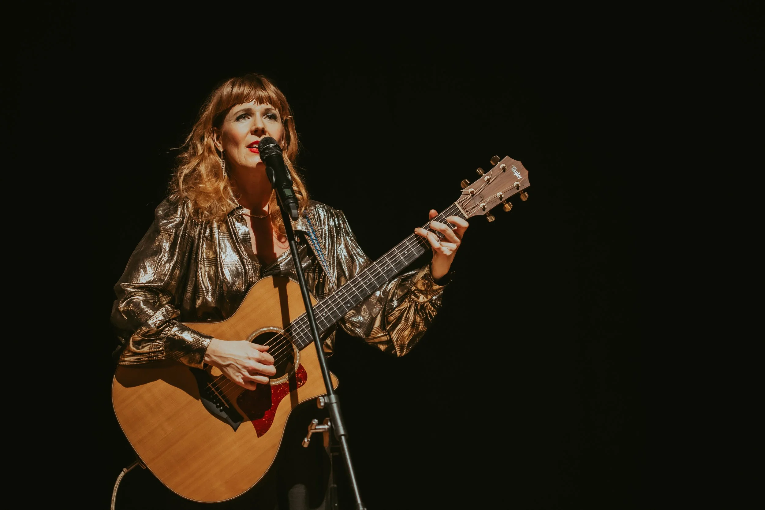 A woman with red hair and a shiny metallic top playing an acoustic guitar on stage, singing into a microphone against a dark background.