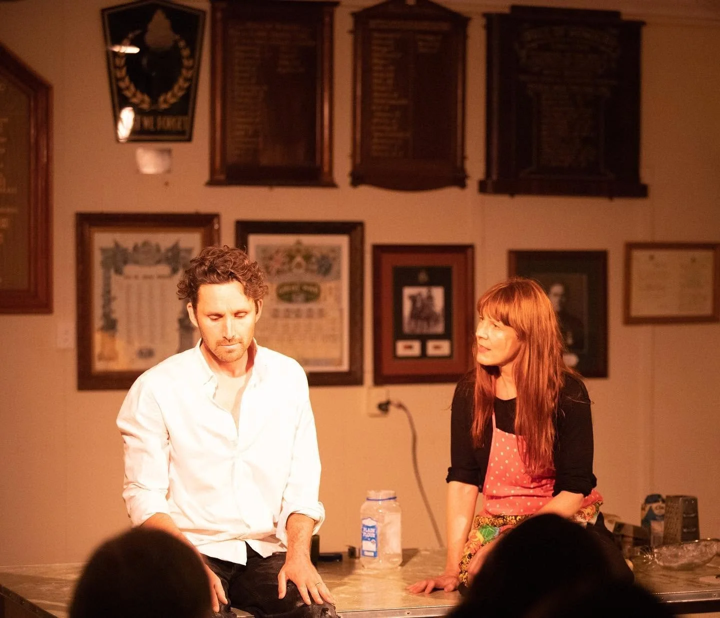 A man and woman sitting on a table in a room decorated with framed pictures and plaques, with some audience members visible in the foreground.