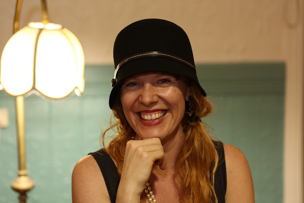 Woman with red hair wearing a black hat, smiling, sitting indoors near a lamp.
