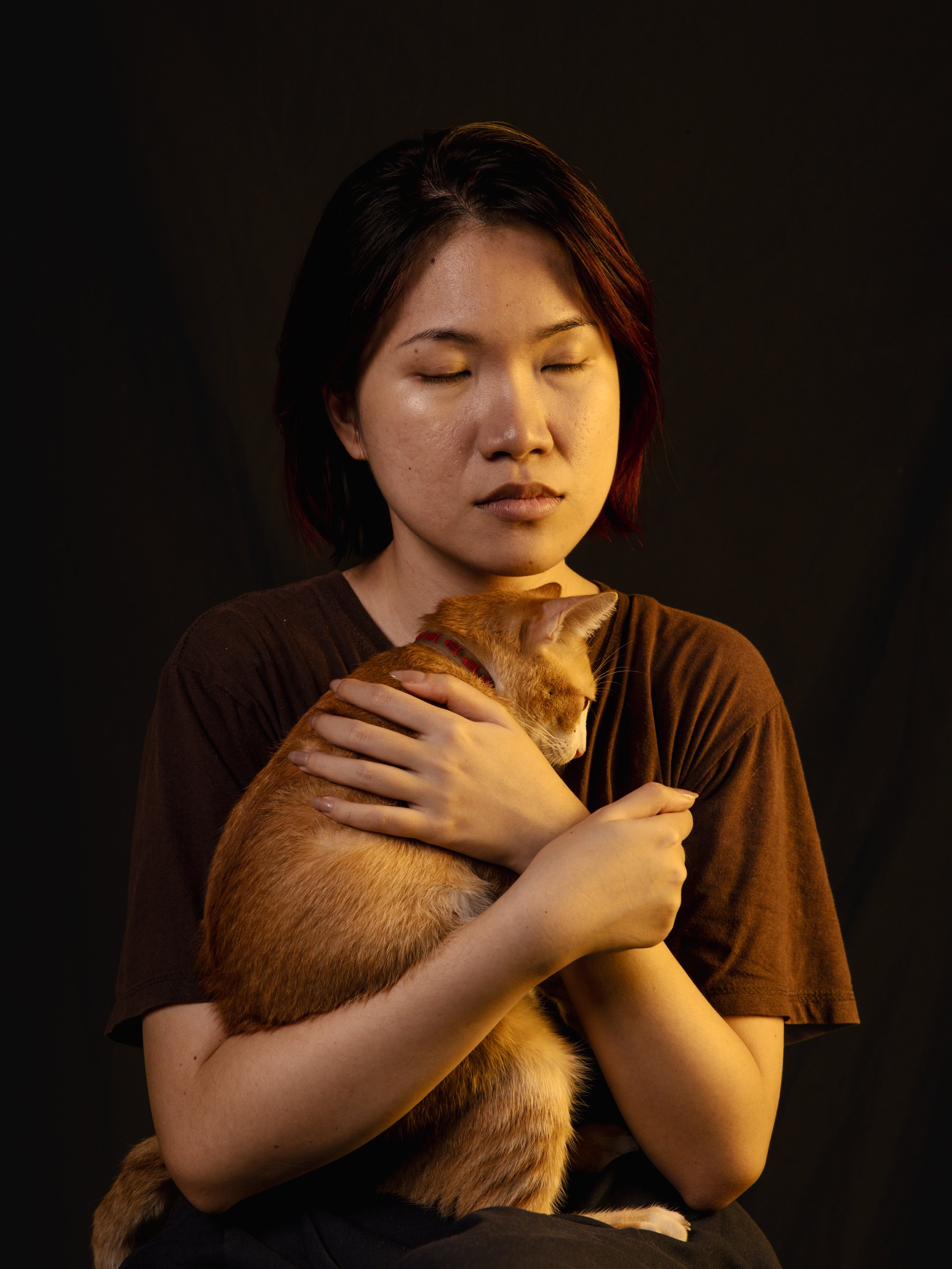 A woman with short dark hair gently holding an orange tabby cat, both appearing calm with eyes closed, against a black background.
